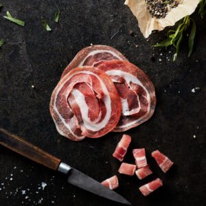 Sliced and diced pancetta on dark table background with wooden handle knife and scattered cracked black pepper