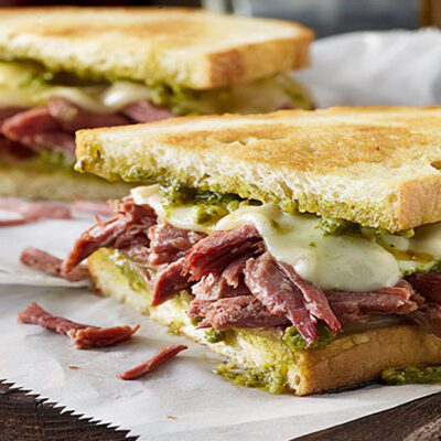 Sandwich, toasted white bread with corned beef pieces, melted cheese and visible pesto