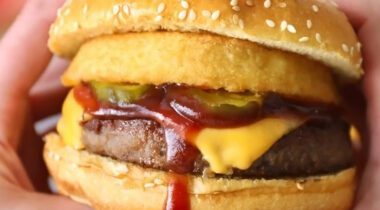 hands holding a cheeseburger with onion rings and pickles on it