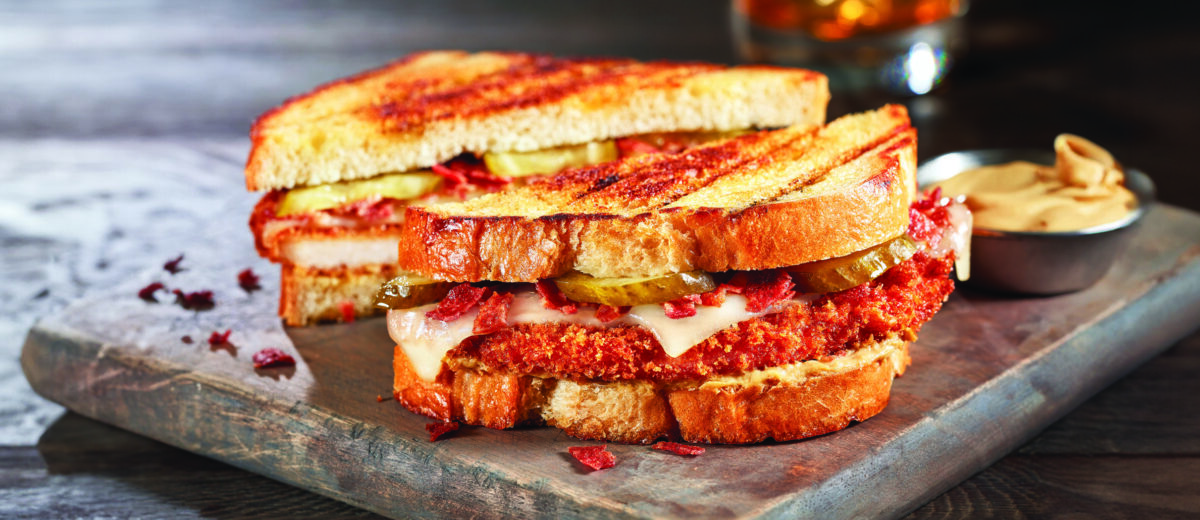 A Turkey Cordon Bleu Sandwihc sitting atop a wooden cutting board, wiht sauce to the right side in a dish, and a beverage in the background