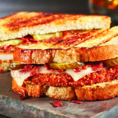 A Turkey Cordon Bleu Sandwihc sitting atop a wooden cutting board, wiht sauce to the right side in a dish, and a beverage in the background