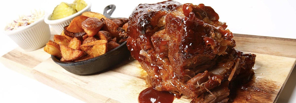 cooked pork butt with bbq sauce on cutting board, with potato wedges in a skillet and coleslaw and pickles in dishes