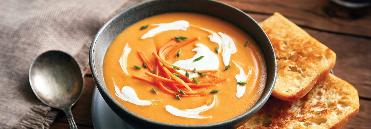 black bowl with orange soup, shaved carrots, and chives. Spoon on left side of bowl with napkin. two pieces of toast on right side of bowl.