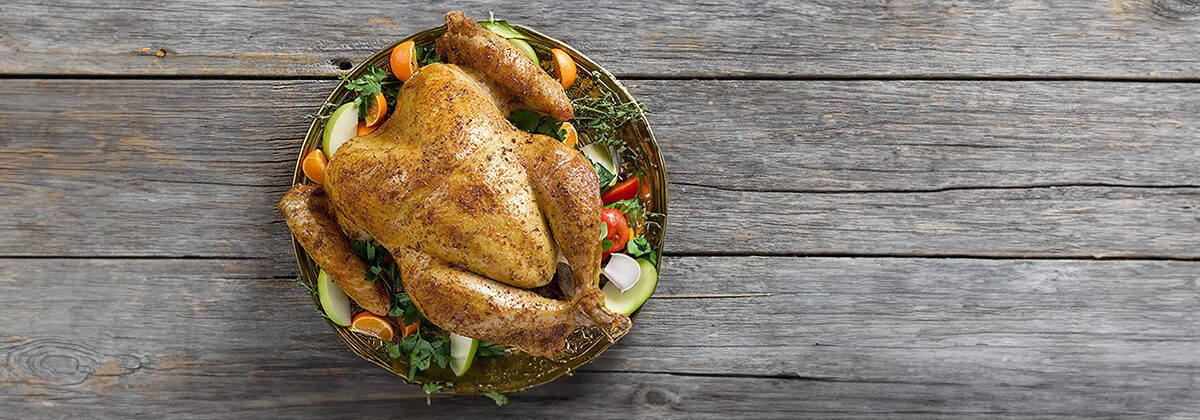 A gray wooden background, with a. big turkey. sitting on a round platter. Surround the turkey on the platter are fresh slices of fruit.